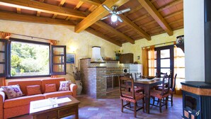 A cozy living room with a wooden ceiling, a fireplace, a dining area with wooden chairs, a sofa with cushions, and a window with curtains. - ClickSardegna Villa Porticciolo near the beach for 6 people, with A/C and WiFi (Alghero)