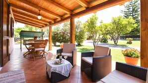 A covered patio with wooden furniture, a table with a bottle and glasses, and a view of a garden with trees and flowers. - ClickSardegna Villa Porticciolo near the beach for 6 people, with A/C and WiFi (Alghero)