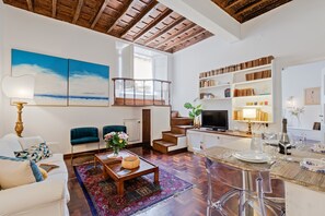 A spacious living area with a wooden ceiling, a large window, a staircase, a dining area, and a bookshelf.