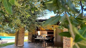 Un espace extérieur aménagé en terrasse, avec une table et des chaises en briques, entouré d’une végétation luxuriante et une piscine en arrière-plan.