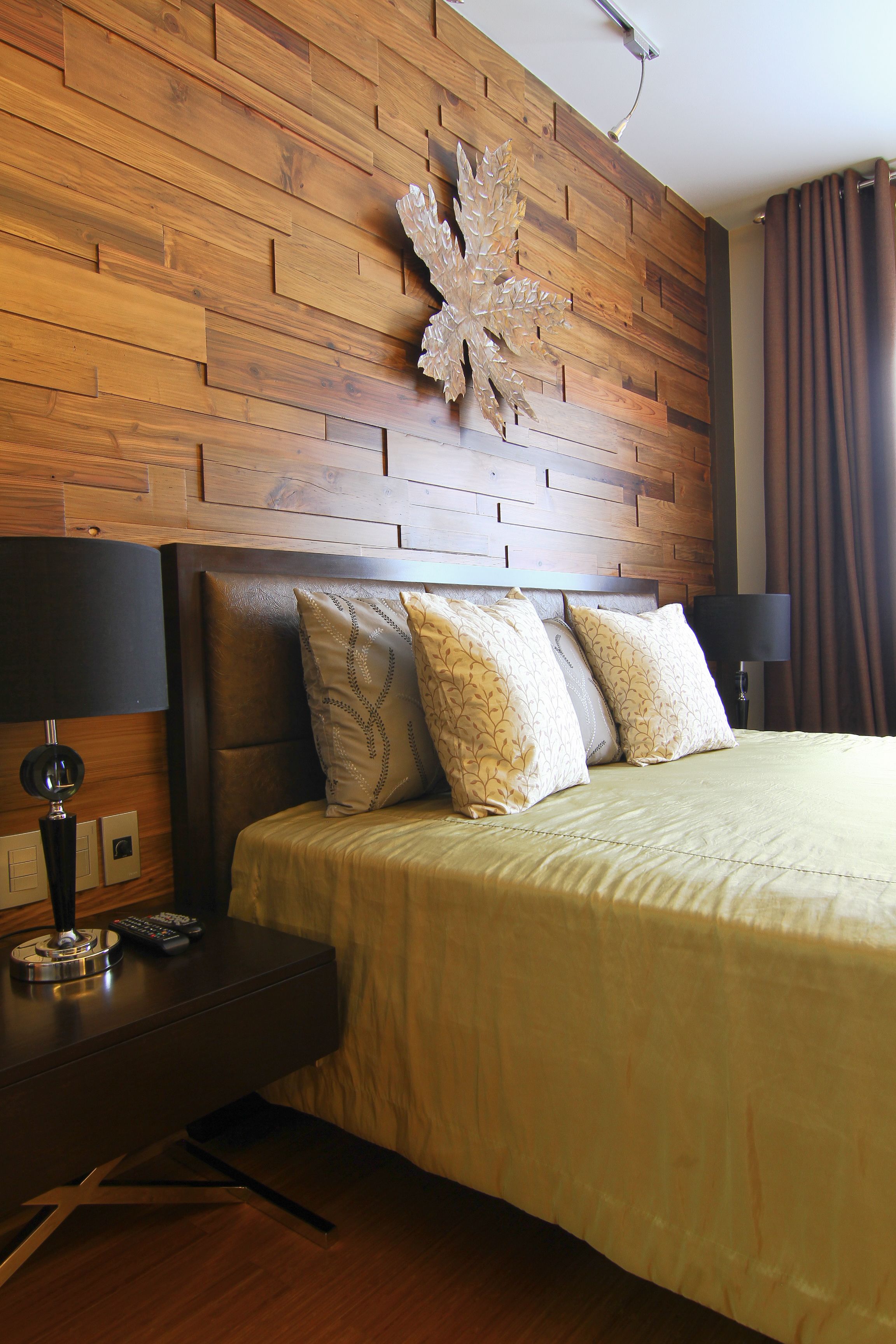Una habitación de hotel con cabecera de madera, cama con varios cojines, mesita de noche con lámpara y control remoto.