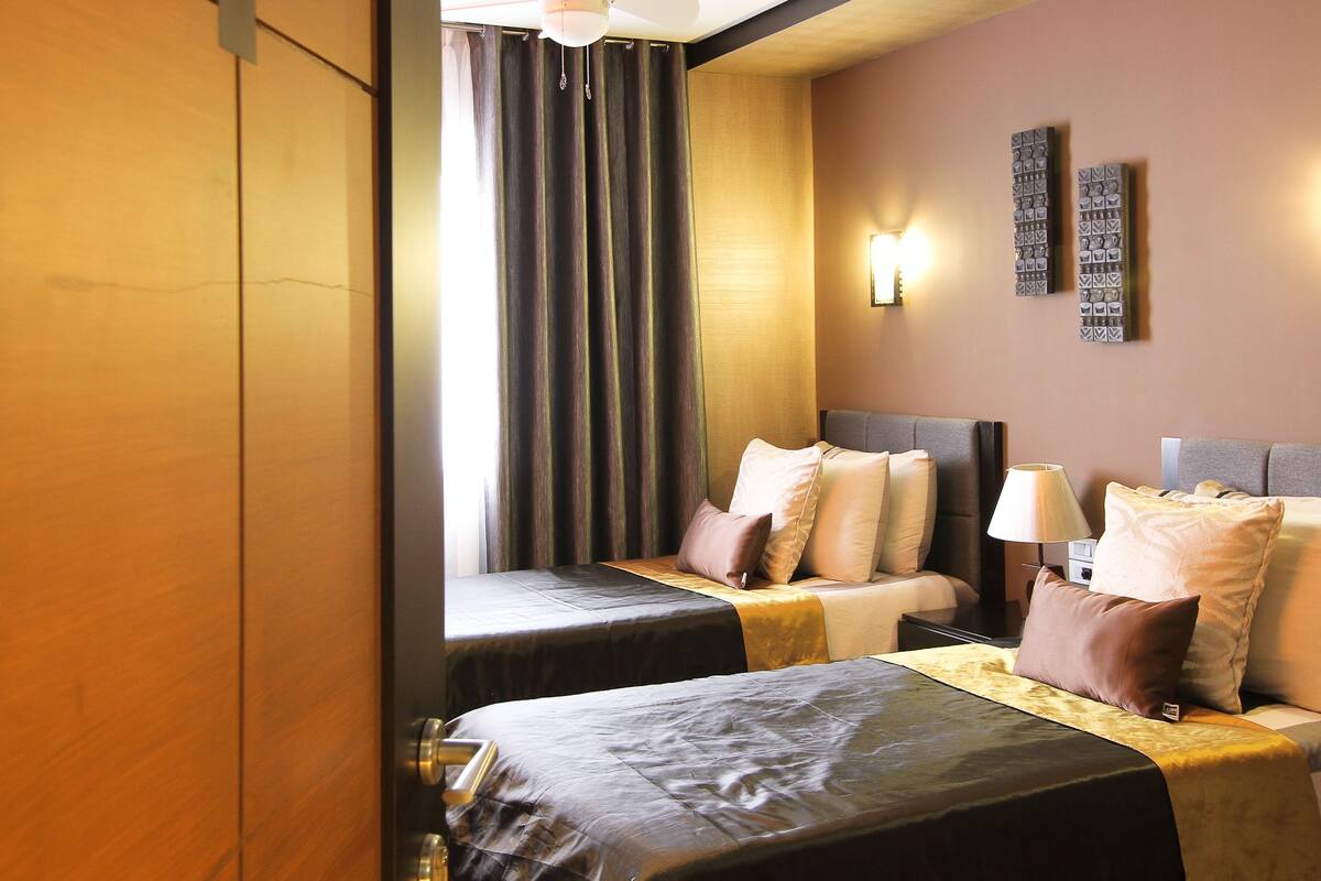 Une chambre d’hôtel avec deux lits, une grande fenêtre avec des rideaux, une lampe fixée au mur et une petite table avec une lampe.