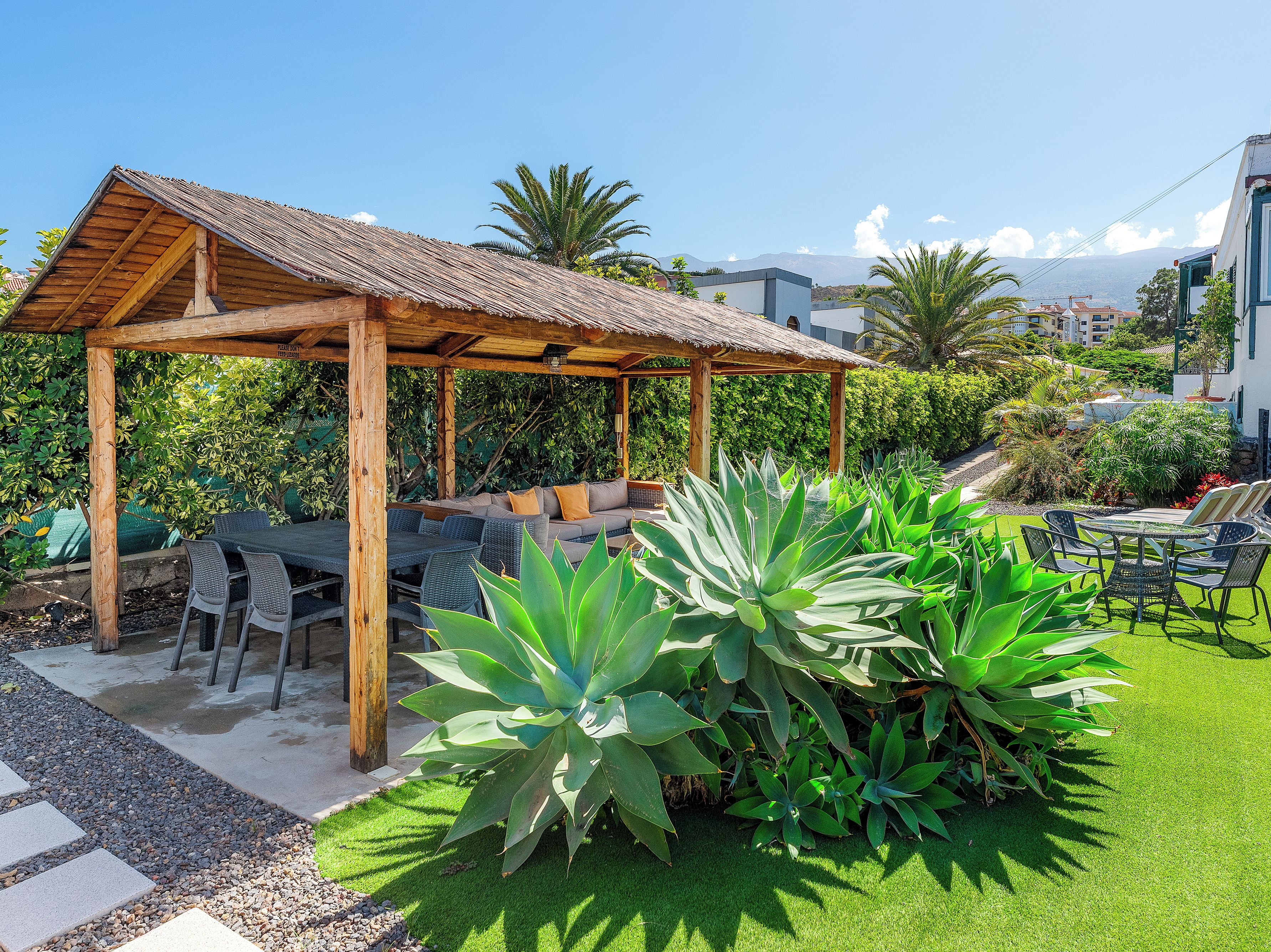 A wooden pergola with outdoor seating, surrounded by lush greenery and a clear sky.