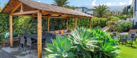 A wooden pergola with outdoor seating, surrounded by lush greenery and a clear sky.