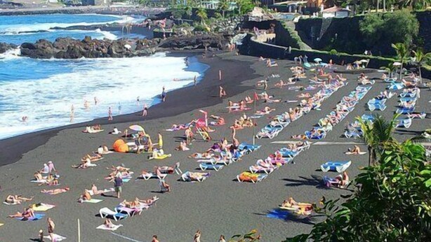 A beach with rows of sun loungers and umbrellas, a rocky shoreline, and buildings in the background.