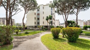 Un immeuble résidentiel à plusieurs étages, accessible par un chemin pavé, entouré d’espaces verts et d’arbres bien entretenus.