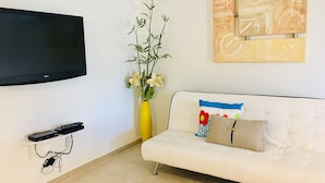 Un salon avec un téléviseur à écran plat fixé au mur, un canapé blanc agrémenté de coussins colorés, un vase jaune avec des fleurs et une petite étagère sur laquelle repose une télécommande.