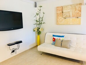 Een woonkamer met een flatscreen televisie aan de muur, een witte bank met kleurrijke kussens, een gele vaas met bloemen, en een klein plankje met een afstandsbediening.
