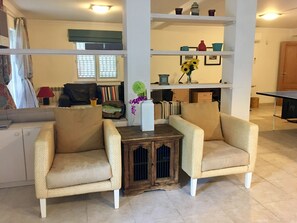 A living room with two beige armchairs, a small wooden table with a vase of flowers, and a bookshelf with various decorative items.