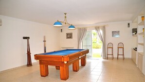 Une table de billard dans une pièce dont le sol est carrelé, avec un ventilateur au plafond et deux tabourets de bar.
