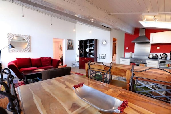 A modern living room with a red sofa, a wooden dining table, and a kitchen area with red walls and stainless steel appliances.