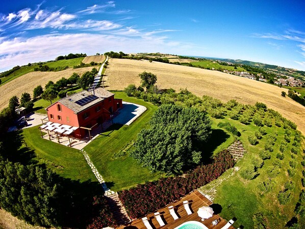 A red building with solar panels, surrounded by green fields and trees.