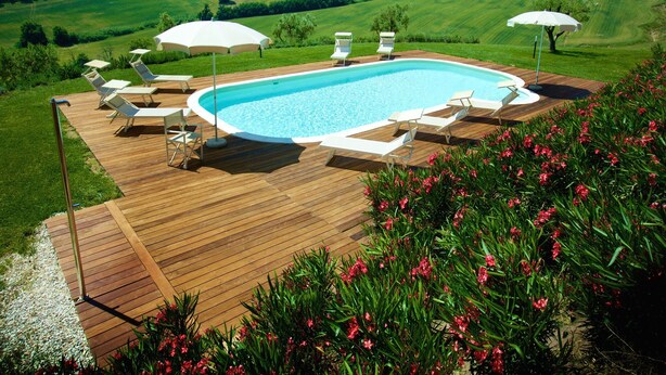 Ein Schwimmbecken mit Holzdeck, umgeben von Liegestühlen und Sonnenschirmen, vor einer malerischen Hügellandschaft.
