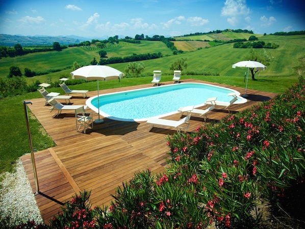 A swimming pool with a wooden deck, surrounded by lounge chairs and umbrellas, set against a scenic hillside backdrop.