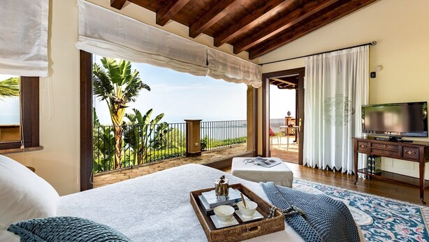 A bedroom with a large bed, a TV, and a balcony with a view of palm trees and the sea. - Villa Alba 12, Emma Villas (Giardini Naxos)