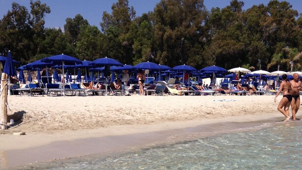 Una playa con aguas transparentes y poco profundas, sombrillas azules y sillas de playa.