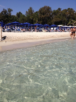 A beach with clear shallow waters, blue umbrellas, and beach chairs.