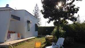 Un edificio blanco con una ventana pequeña y una puerta, un patio cubierto de césped y dos sillas blancas.