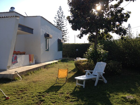 A white building with a small window and a door, a grassy yard, and two white chairs.