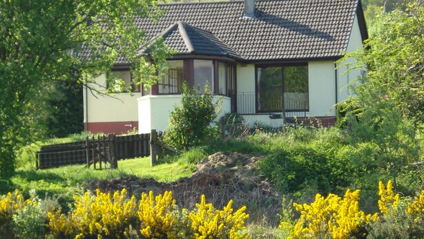 A house with a tiled roof, a fenced yard, and a river in the foreground.