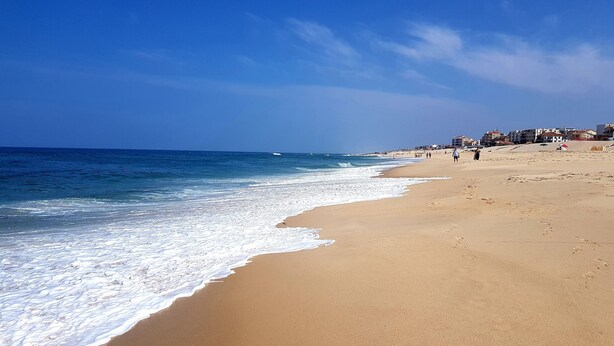 A wide sandy beach with gentle waves, a clear blue sky, and buildings in the background. - T2 by the beach, with pool, WIFI and cable TV, in Praia do Pedrogão (Coimbrão)