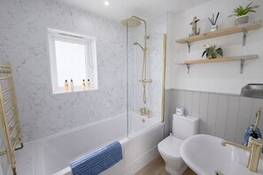 A bathroom with a bathtub, toilet, and shower. There are shelves with toiletries and a window above the bathtub. - Escape to Beach Boutique: A Luxurious Haven in Camber Sands (Camber)