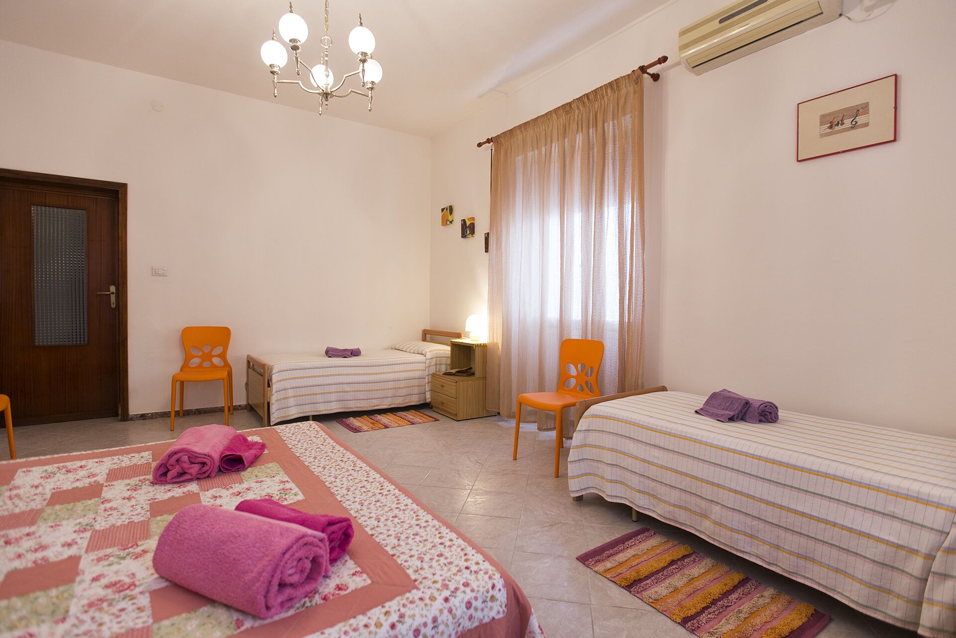 A hotel room with two single beds, a wooden door, a chandelier, a window with curtains, and a wall-mounted air conditioner.