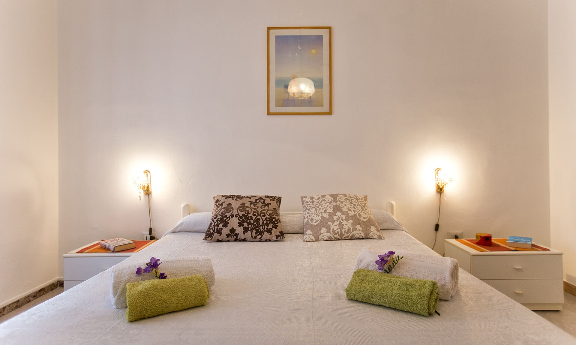 Una cama bien hecha con dos almohadas, dos toallas enrolladas y un cuadro en la pared.