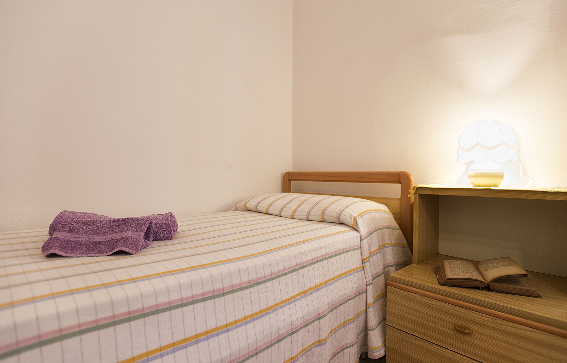 Una cama individual con una colcha a rayas, una toalla morada, un cabecero de madera, una mesita de noche con una lámpara y un atril con un libro abierto.