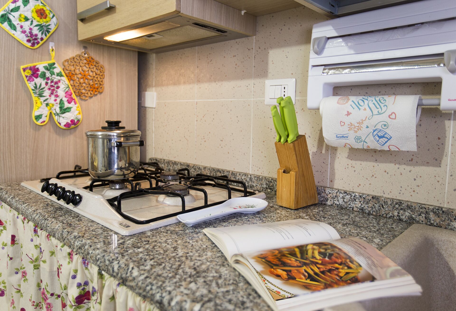 A kitchen with a gas stove, a pot on the stove, a knife block with green handles, and a cookbook on the counter.