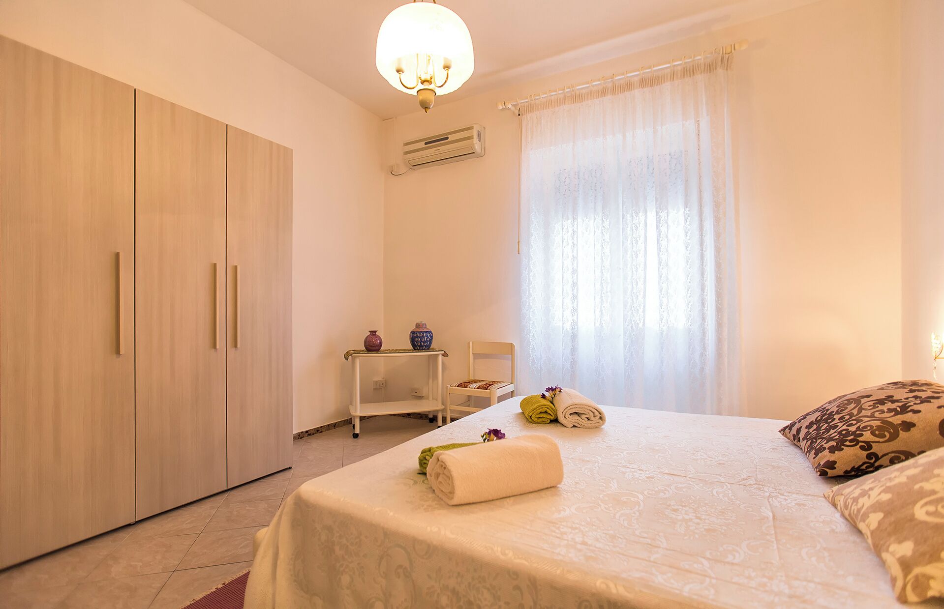 A bedroom with a bed, a wardrobe, a small table with a vase, and a chair.