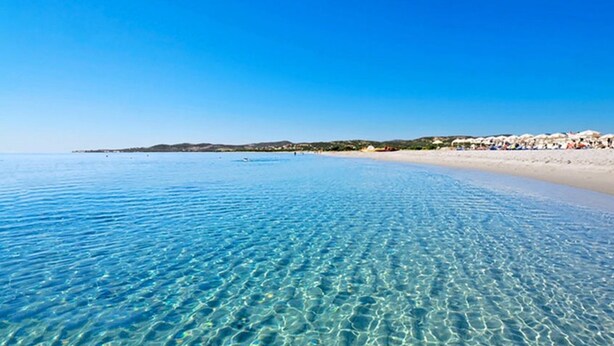 A clear beach with shallow water, a sandy shore, and a distant shoreline with buildings and umbrellas. - Alghero Apartment Cervi 30 meters from Alghero beach for 6 persons (Alghero)