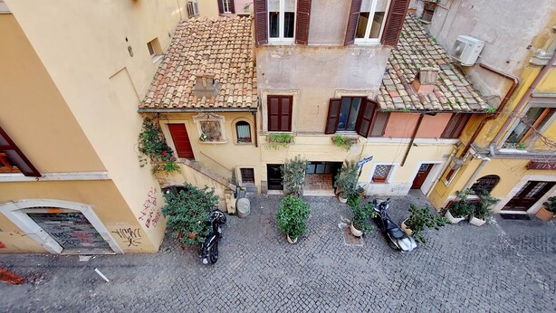 A narrow cobblestone street with buildings on either side, featuring balconies with potted plants and a few parked scooters.