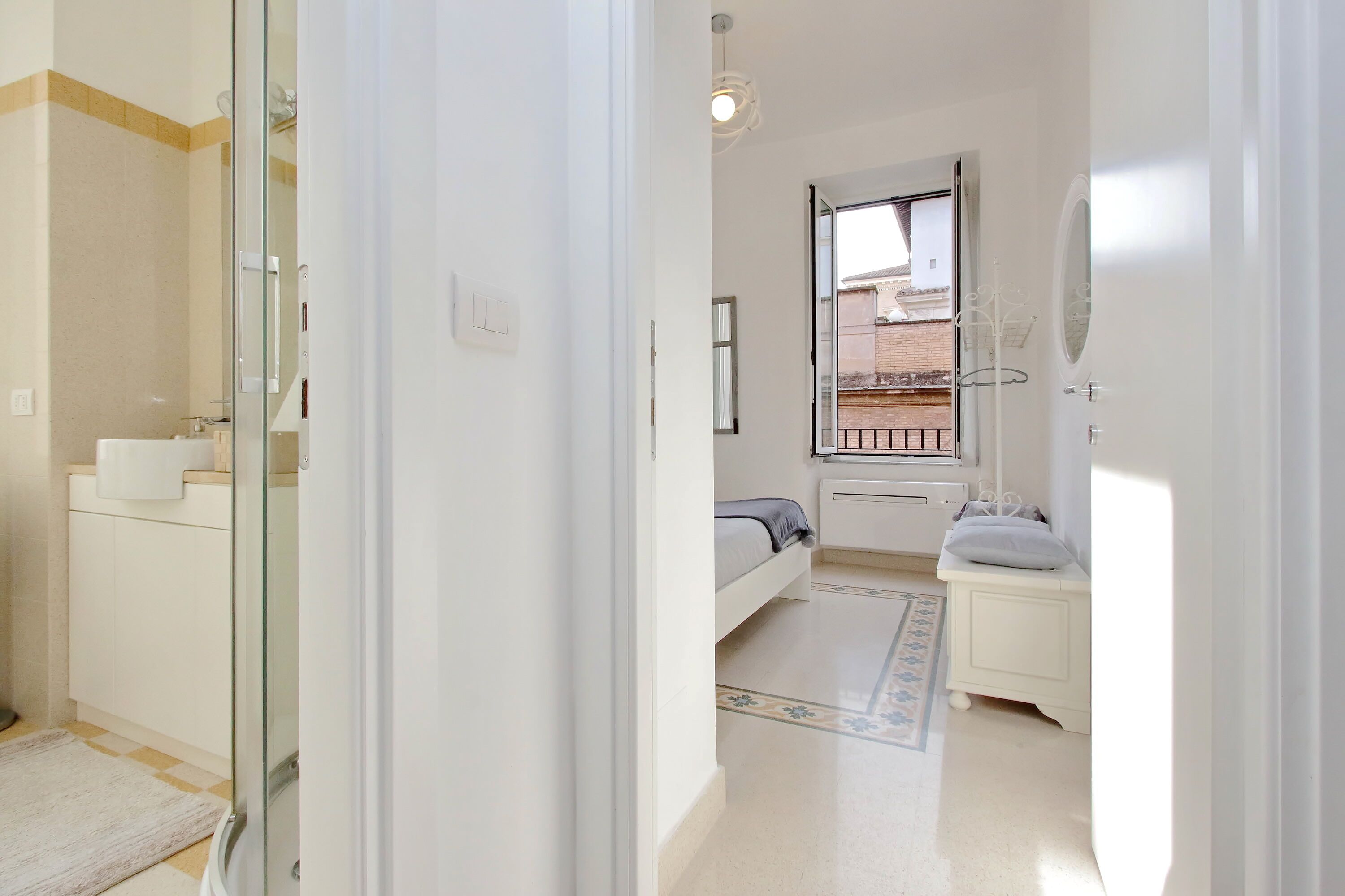 A bedroom with a bed, a bedside table, a window, and a bathroom with a toilet and sink.