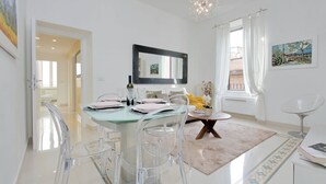 A modern dining area with a glass table, clear chairs, a chandelier, and a painting on the wall.