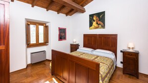 Un dormitori amb sòls de fusta, un llit amb estampa, una tauleta de nit, un radiador, una finestra i un quadre a la paret.