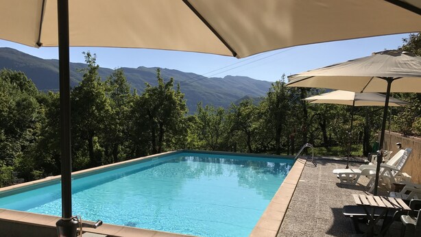 Una piscina rectangular amb aigua transparent de color blau, envoltada per un solàrium amb mosaics de petits pedres. Hi ha gandules i ombrelles que ofereixen ombra, i la piscina fa de fons a arbres verds i muntanyes llunyanes.