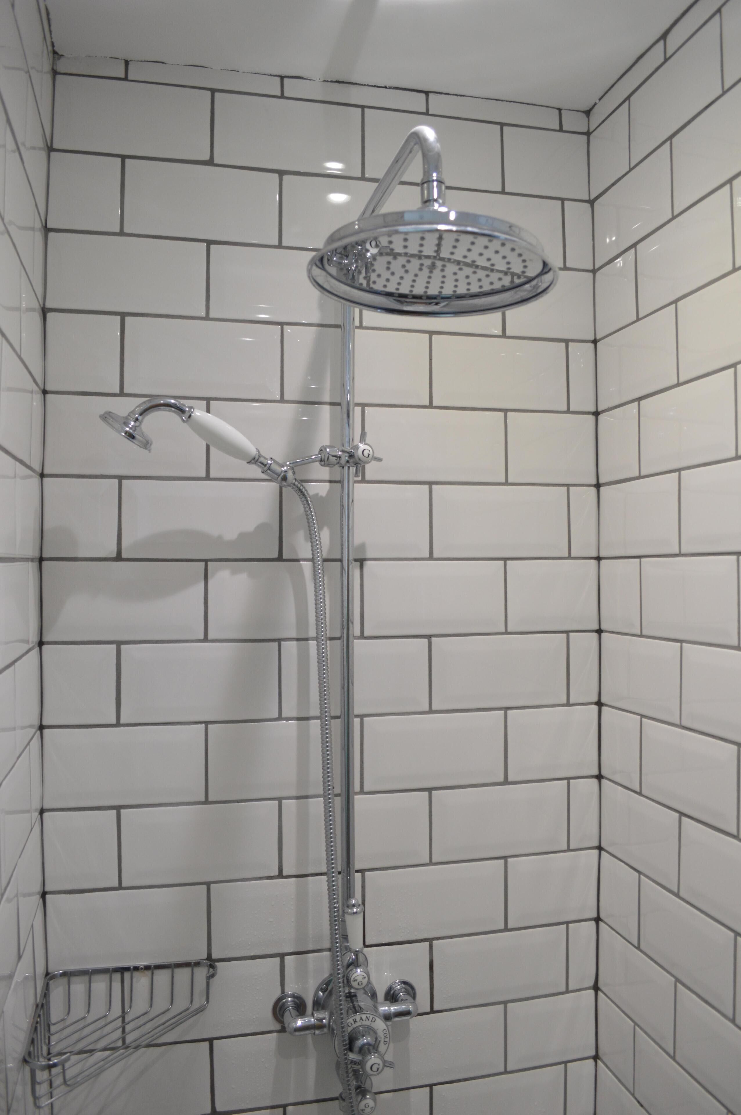 Doccia con soffione a mano e pioggia verticale integrati, entrambi con finiture cromate, in un bagno con pareti rivestite di piastrelle bianche a metro.