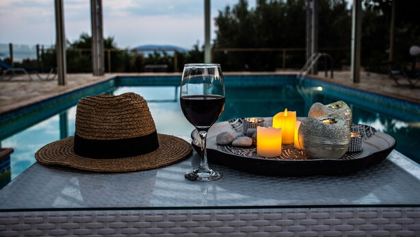 Un verre de vin rouge, un chapeau de paille et des objets décoratifs sur une table près d’une piscine.