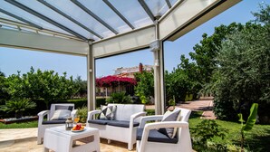 Une terrasse extérieure avec des meubles en osier blanc, une table en verre et une vue sur un jardin et un bâtiment en arrière-plan.