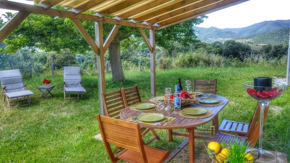 Un espace extérieur aménagé pour les repas, avec une pergola en bois, une table ronde dressée pour quatre personnes et une vue sur un paysage montagneux en arrière-plan.