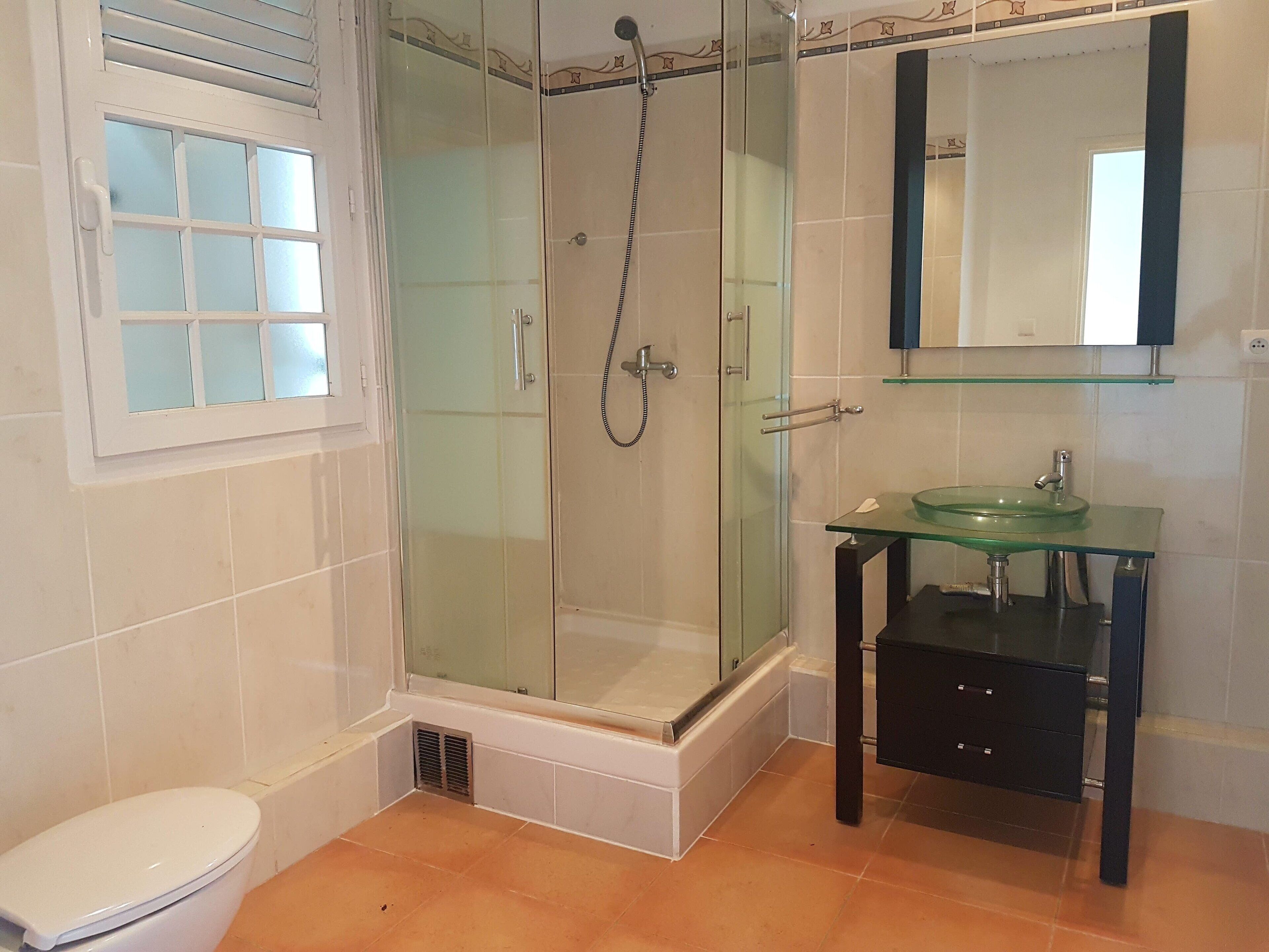 A bathroom with a glass-enclosed shower, a toilet, and a glass sink with a black cabinet.