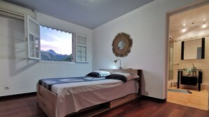 A bedroom with a bed, a window with shutters, a mirror, and a bathroom with a sink and mirror.