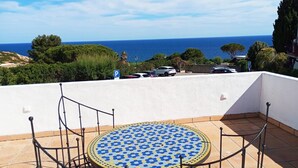 Una terraza con mesa y sillas de cerámica, con vistas a una zona costera con coches aparcados y cielo despejado.