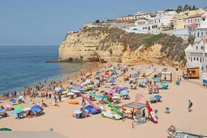 Praia cheia, com guarda-sóis coloridos e um escorrega, com vista para um penhasco e edifícios.