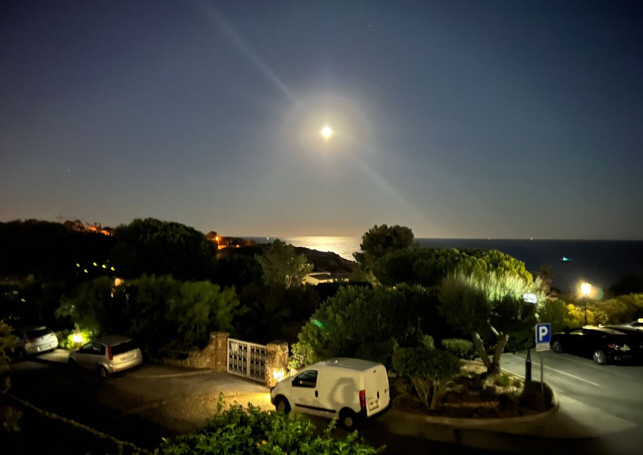 A nighttime view of a coastal area with parked cars and a white van, a parking sign, and a distant view of the sea.