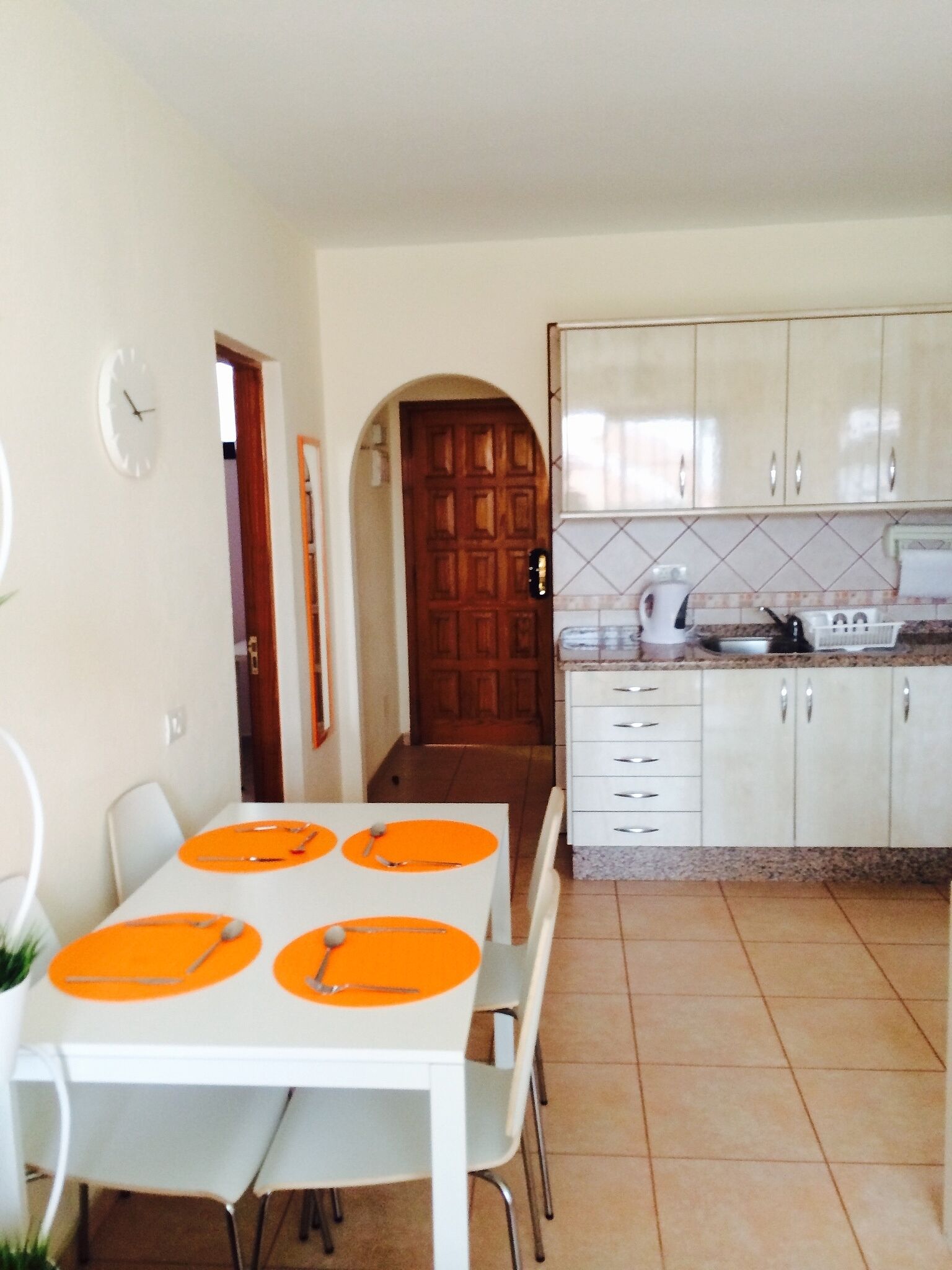 Comedor con una mesa blanca para cuatro, cocina con armarios blancos y salpicadero de azulejos, y puerta de madera que da a otra habitación.
