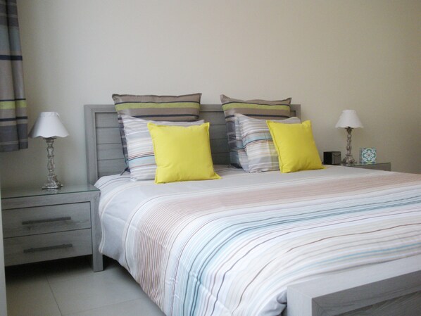 Una cama bien hecha con ropa de cama a rayas y dos almohadas amarillas, una mesita de noche con una lámpara y otra mesita de noche también con una lámpara.