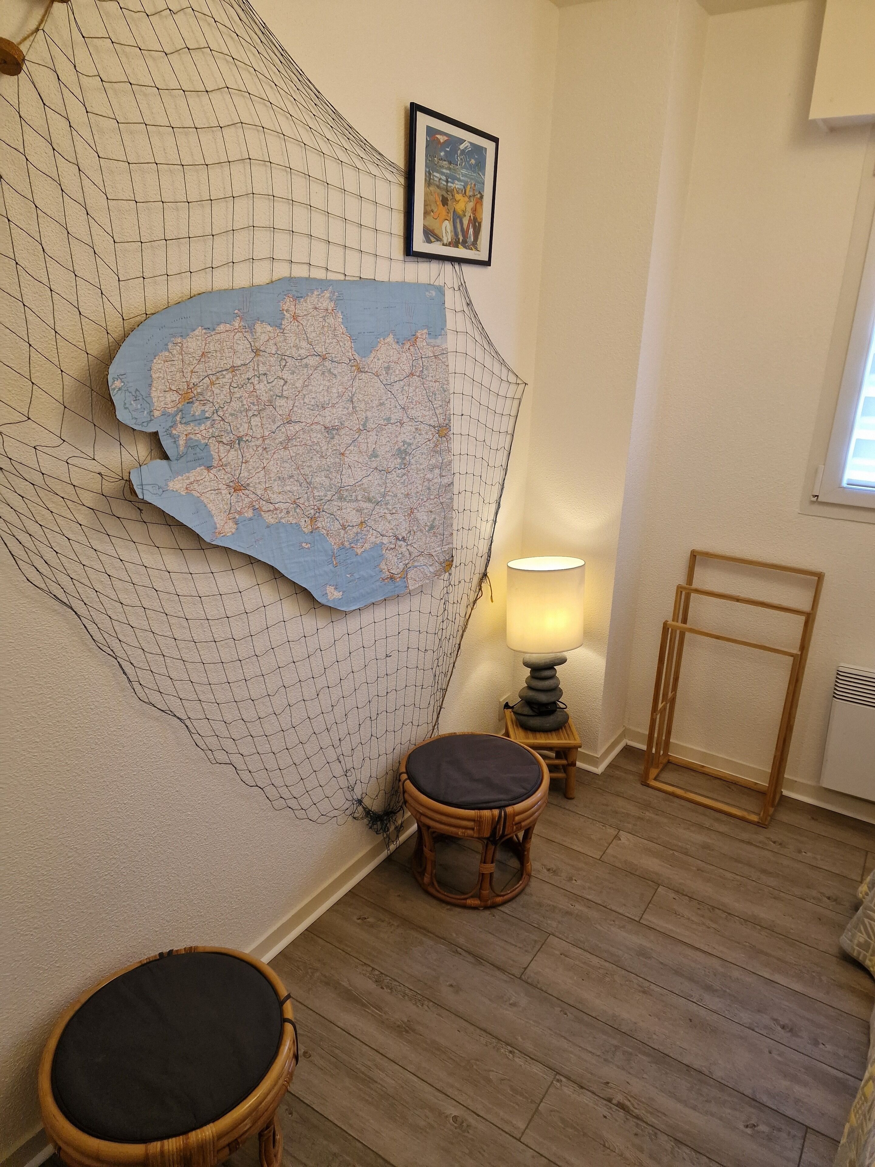 Une pièce avec une grande carte accrochée au mur, une petite table ronde avec un coussin, un lampadaire et un cadre en bois appuyé contre le mur.