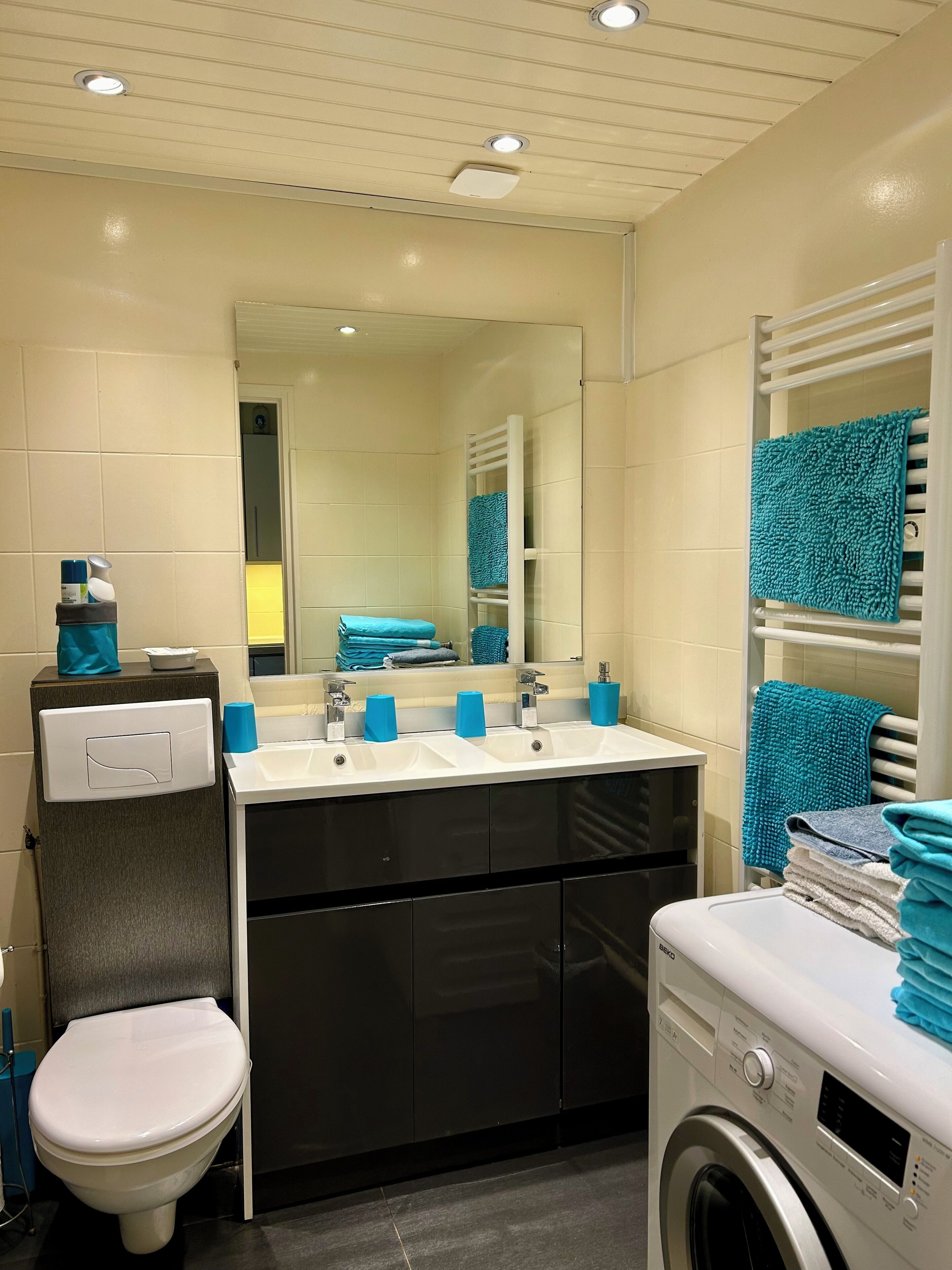 Un baño moderno con inodoro blanco, un lavamanos con dispensadores de jabón azules, un espejo y una lavadora.